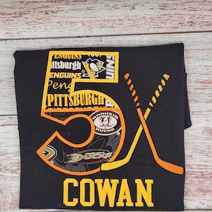 May include: Black t-shirt with an embroidered hockey-themed design. The design includes the number "5X" in orange and yellow, crossed hockey sticks, and the name "COWAN" in yellow. Logos for the Pittsburgh Penguins and Anaheim Ducks are also featured.