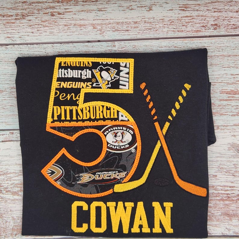 May include: Black t-shirt with an embroidered hockey-themed design. The design includes the number "5X" in orange and yellow, crossed hockey sticks, and the name "COWAN" in yellow. Logos for the Pittsburgh Penguins and Anaheim Ducks are also featured.