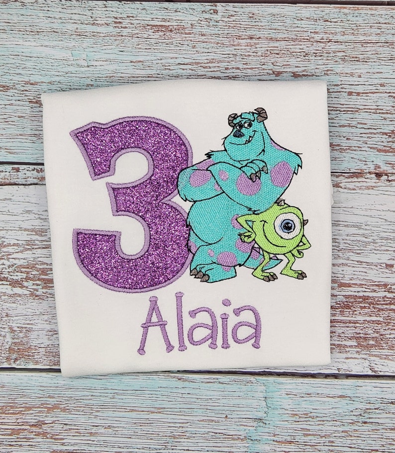 May include: Purple glitter number 3 with embroidered images of Sully and Mike from Monsters Inc. The name Alaia is embroidered below the number.