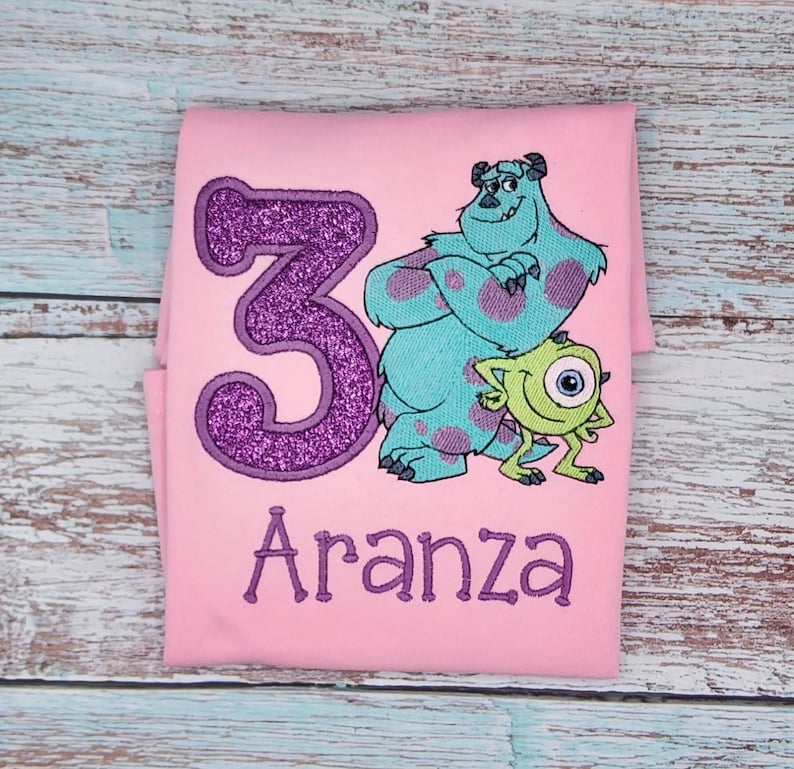 May include: Pink t-shirt with a purple glittery number 3, a blue monster with purple spots, a green one-eyed monster, and the name "Aranza" embroidered in purple.