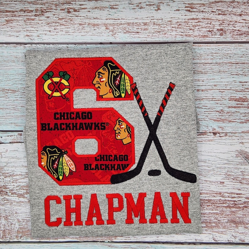 May include: Gray t-shirt featuring a red number 6 with the Chicago Blackhawks logo and the name "CHAPMAN" in red. Two black hockey sticks are crossed behind the number.