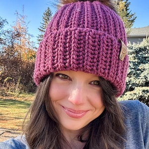 Könnte beinhalten: Nahaufnahme einer gestrickten magentafarbenen Mütze mit einem großen braunen Kunstfell-Bommel oben. Die Mütze hat ein geripptes Design und ein kleines Lederetikett. Der Hintergrund zeigt einen blauen Himmel und Bäume.