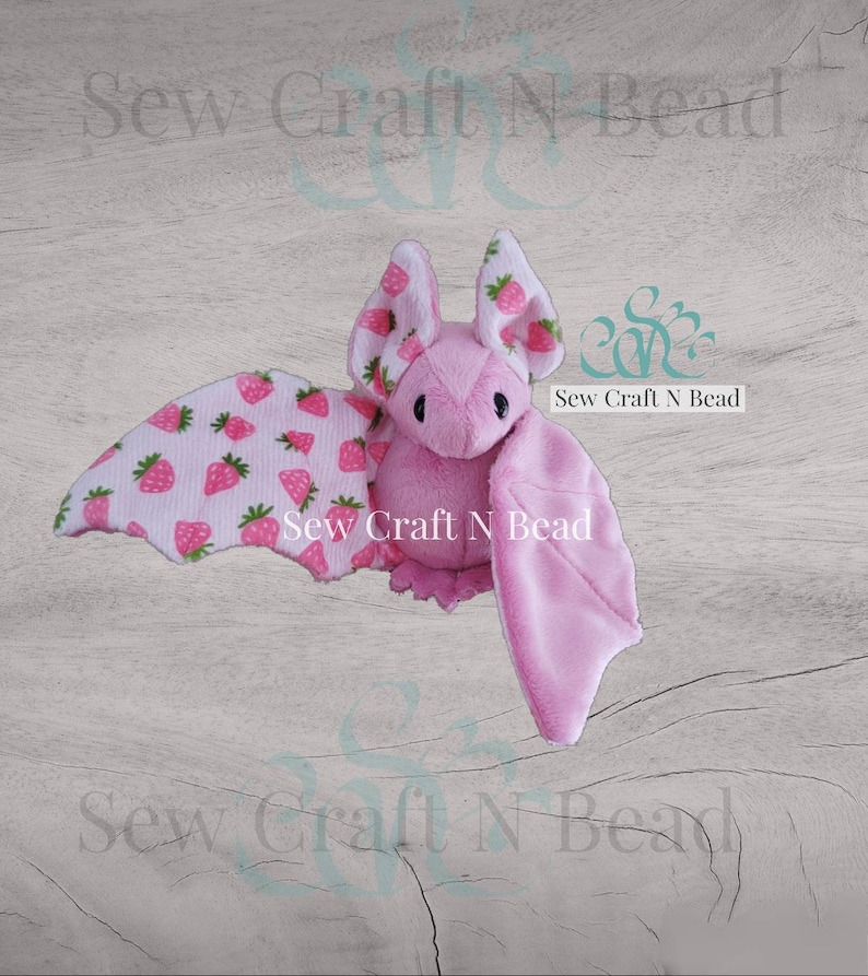 Puede incluir: Murci&eacute;lago de peluche rosa con alas de estampado de fresa. El murci&eacute;lago tiene un cuerpo rosa y ojos negros. El texto "Sew Craft N Bead" est&aacute; impreso en las alas.