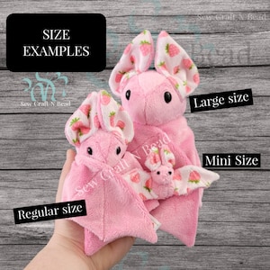 Puede incluir: Tres murci&eacute;lagos de peluche rosa de diferentes tama&ntilde;os con alas de tela de estampado de fresa. Los murci&eacute;lagos est&aacute;n etiquetados como "Tama&ntilde;o grande", "Tama&ntilde;o regular" y "Tama&ntilde;o mini".
