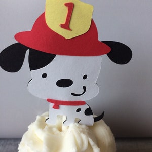 May include: A white and black dalmatian dog wearing a red firefighter hat with a yellow badge that says "1" on it. The dog is sitting on top of a white frosted cupcake.