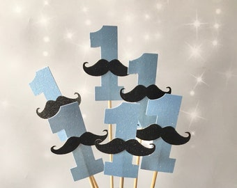 May include: Six blue glitter paper number one cutouts with black paper moustaches attached to wooden skewers. The skewers are inserted into a clear glass jar filled with silver glitter balls.