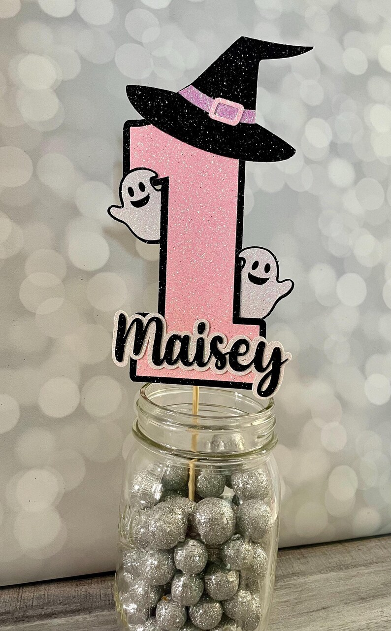 May include: A pink glittery number one cake topper with a black witch hat and two ghost cutouts. The name "Maisey" is written in black and white script. The topper is in a jar filled with silver glitter balls.