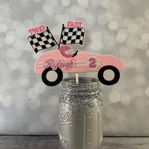 May include: Pink race car cake topper with black and white checkered flags, the words "Two Fast" and the name "Ryleigh" with the number "2" on the side.