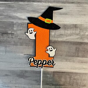 May include: A Halloween-themed cake topper featuring a large orange number one with a black witch hat and two white ghost figures. The name "Pepper" is written in a cursive font at the bottom. The topper is attached to a white stick.