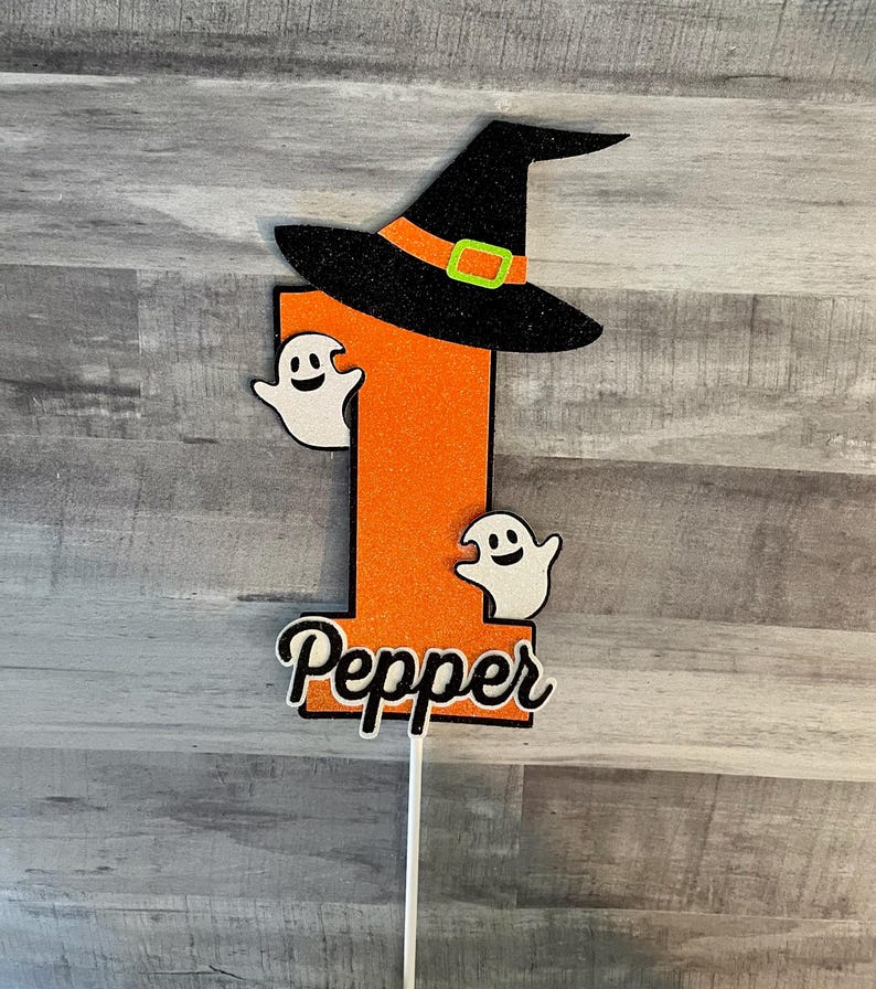 May include: A Halloween-themed cake topper featuring a large orange number one with a black witch hat and two white ghost figures. The name "Pepper" is written in a cursive font at the bottom. The topper is attached to a white stick.
