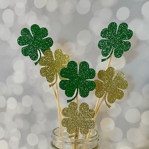 May include: A set of six green and gold glitter shamrock picks. The picks are arranged in a mason jar with silver beads at the bottom.