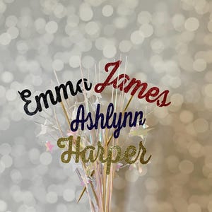 May include: A bouquet of glittery name toppers on wooden sticks. The names Emma, James, Ashlynn, and Harper are displayed in black, red, blue, and gold glitter, respectively. The arrangement includes star-shaped accents and a soft, blurred background.