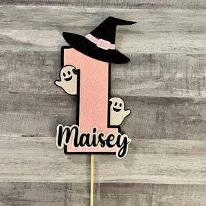 May include: A pink glittery number one cake topper with a black witch hat and two smiling ghost cutouts. The name "Maisey" is written in cursive below the number. The topper is on a wooden stick.