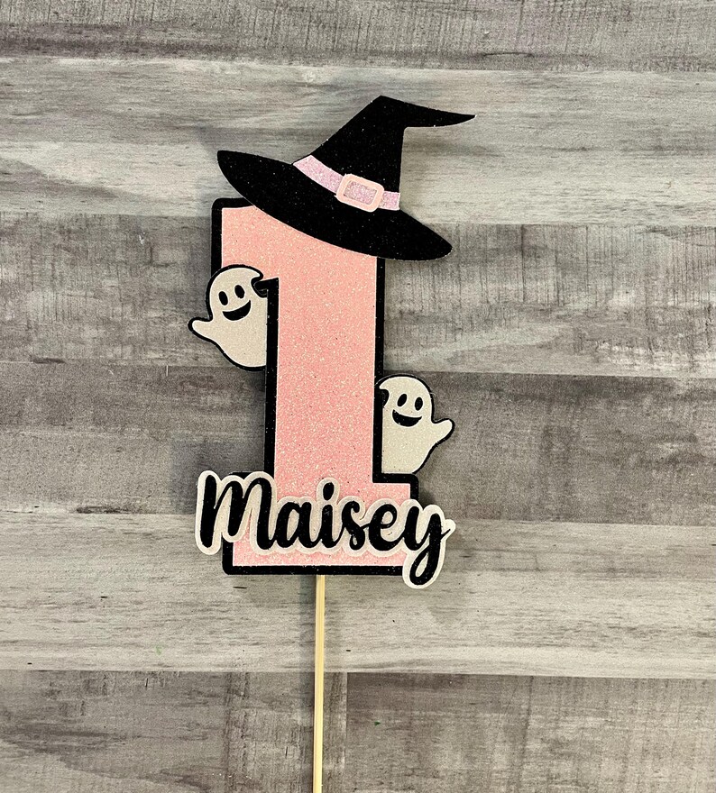May include: A pink glittery number one cake topper with a black witch hat and two smiling ghost cutouts. The name "Maisey" is written in cursive below the number. The topper is on a wooden stick.