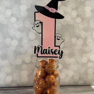 May include: A Halloween-themed birthday cake topper featuring a pink glitter number one, a black witch hat, and two smiling ghost cutouts. The name "Maisey" is displayed below the number. The topper is placed in a glass jar filled with orange glitter balls.