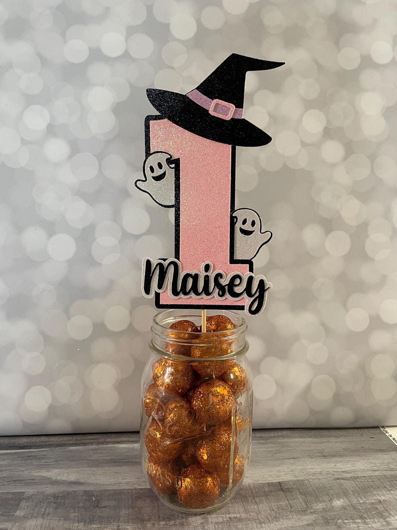 May include: A Halloween-themed birthday cake topper featuring a pink glitter number one, a black witch hat, and two smiling ghost cutouts. The name "Maisey" is displayed below the number. The topper is placed in a glass jar filled with orange glitter balls.