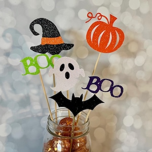 May include: Halloween-themed decorations in a glass jar. The decorations include a witch hat, pumpkin, ghost, bat, and the word "BOO" in green and purple glitter. The decorations are on wooden sticks.
