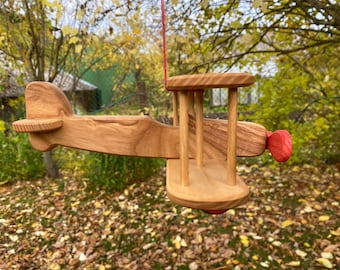 May include: A wooden toy airplane with red accents. The plane has a propeller and a cockpit. The toy is hanging from a red string.