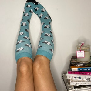 May include: Teal knee-high socks featuring a repeating pattern of black and white dog faces. The socks, with black tops, are worn on legs. A stack of books and a candle are visible in the background.