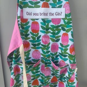 May include: Floral apron featuring pink and orange flowers and green leaves on a light blue backdrop. The apron has a pink pocket and trim, with the text "Did you bring the Gin?" displayed on the front.