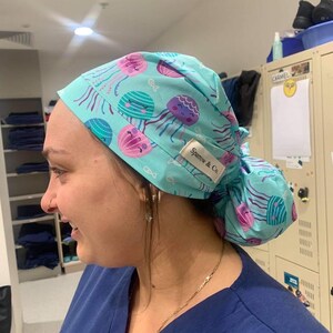 May include: A light blue scrub cap featuring a pattern of pink and teal jellyfish. The cap has a gathered back and a small white tag that reads "Sparrow & Co."