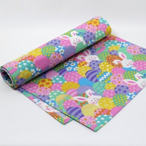 May include: A pink and purple table runner with a pattern of colorful Easter eggs and white bunnies. The runner is rolled up on one end.