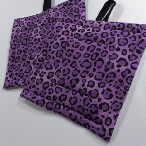 May include: Two square purple potholders with a black leopard print pattern. Each potholder has a black fabric loop for hanging. The potholders are quilted with a stitched border.