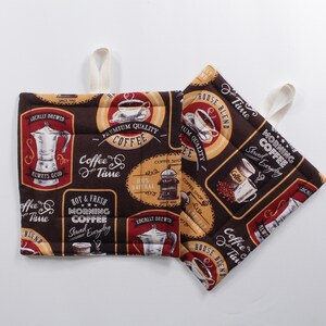May include: Two brown quilted pot holders with a coffee theme. One pot holder features a coffee grinder, a coffee pot, and the text "Coffee Time". The other pot holder features a coffee cup, a coffee pot, and the text "Morning Coffee Served Everyday".