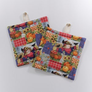 May include: Two quilted pot holders with a patchwork design featuring scarecrows, pumpkins, sunflowers, and plaid patterns in shades of red, blue, yellow, and green.