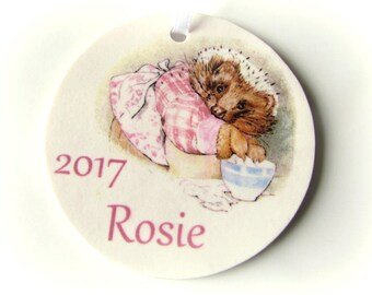 Op de afbeelding: Een ronde, crèmekleurige ornament met een aquarel illustratie van een egel met een muts en schort, met de naam "Rosie" en het jaar "2017" in roze gedrukt. Een wit lint is bevestigd om op te hangen.