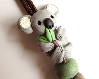 Op de afbeelding: Een handgemaakte koala beer hanger met een bruin koord. De grijze koala houdt een groen blad en een bruine tak vast. De hanger heeft een reeks groene en bruine kralen. Het koord is een gevlochten bruin.