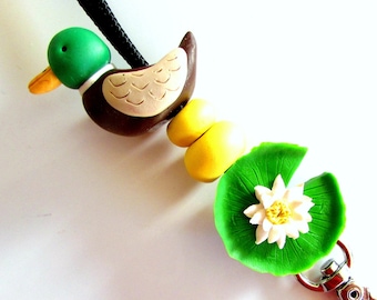 May include: A green and brown duck charm with yellow and white beads and a green lily pad charm attached to a silver key chain.