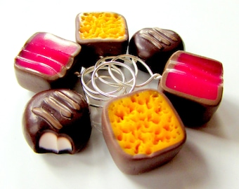 May include: Six chocolate candies with different fillings. The candies are shaped like squares and circles. The fillings are yellow, pink, and white. The candies are arranged in a circle.