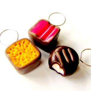 May include: Three chocolate candies with different fillings, each attached to a silver ring. The candies are shaped like squares and a dome. The fillings are pink, yellow, and white.