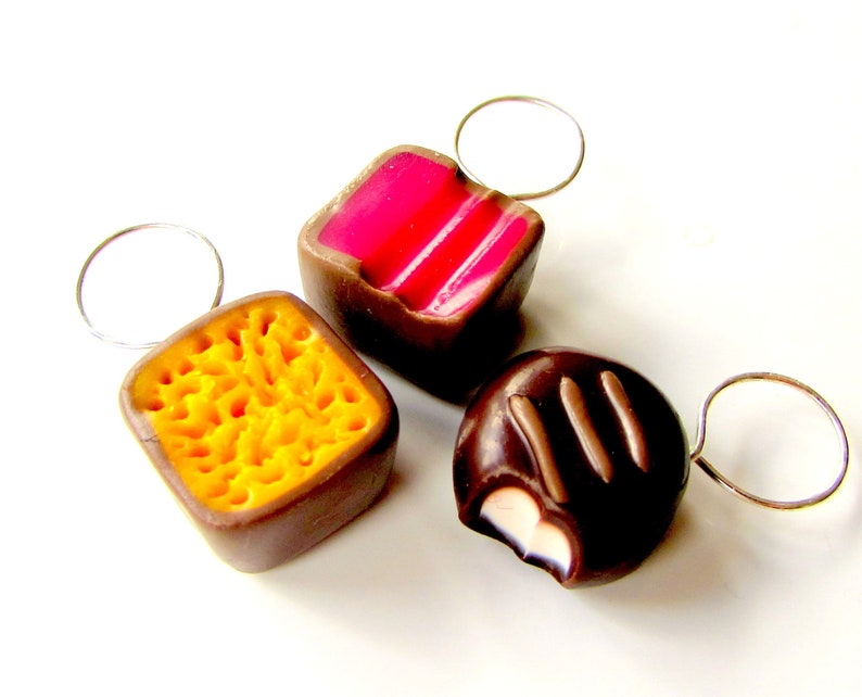 May include: Three chocolate candies with different fillings, each attached to a silver ring. The candies are shaped like squares and a dome. The fillings are pink, yellow, and white.