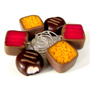 May include: A set of six chocolate candies, each with a different filling. The candies are arranged in a circle and are connected by silver rings. The candies are brown, yellow, and pink.