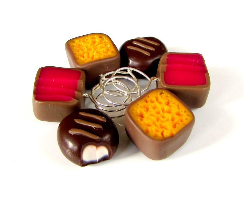May include: A set of six chocolate candies, each with a different filling. The candies are arranged in a circle and are connected by silver rings. The candies are brown, yellow, and pink.