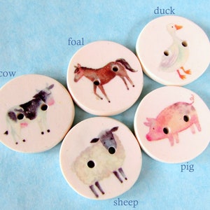 May include: Five round, white, ceramic buttons with different animal illustrations. The buttons feature a cow, a foal, a duck, a pig, and a sheep.