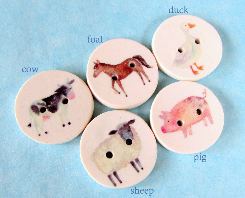 May include: Five round, white, ceramic buttons with different animal illustrations. The buttons feature a cow, a foal, a duck, a pig, and a sheep.