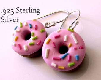 May include: A pair of .925 Sterling Silver donut earrings. Each pink donut is decorated with colorful sprinkles and has a brown base. The earrings have silver hooks.