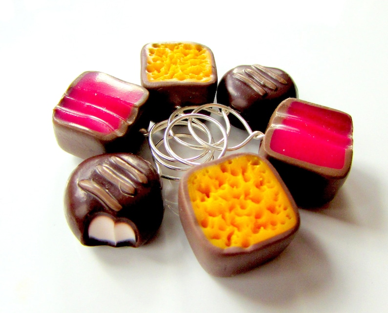 May include: Six chocolate candies with different fillings. The candies are shaped like squares and circles. The fillings are yellow, pink, and white. The candies are arranged in a circle.
