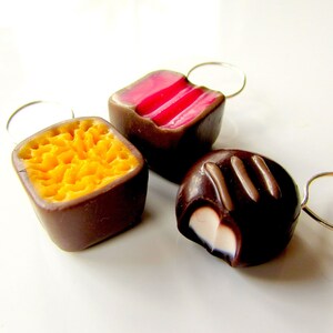 May include: Three chocolate candies with different fillings. The first candy is a square with a yellow honeycomb filling. The second candy is a square with a pink and white striped filling. The third candy is a round candy with a white filling and chocolate topping.
