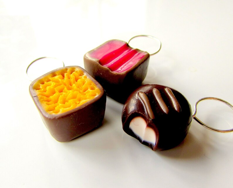 May include: Three chocolate candies with different fillings. The first candy is a square with a yellow honeycomb filling. The second candy is a square with a pink and white striped filling. The third candy is a round candy with a white filling and chocolate topping.