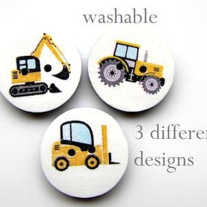 May include: Three round, white buttons with yellow construction vehicles printed on them. The buttons feature a yellow excavator, a yellow tractor, and a yellow forklift. The text "washable" and "3 different designs" is printed on the white background.