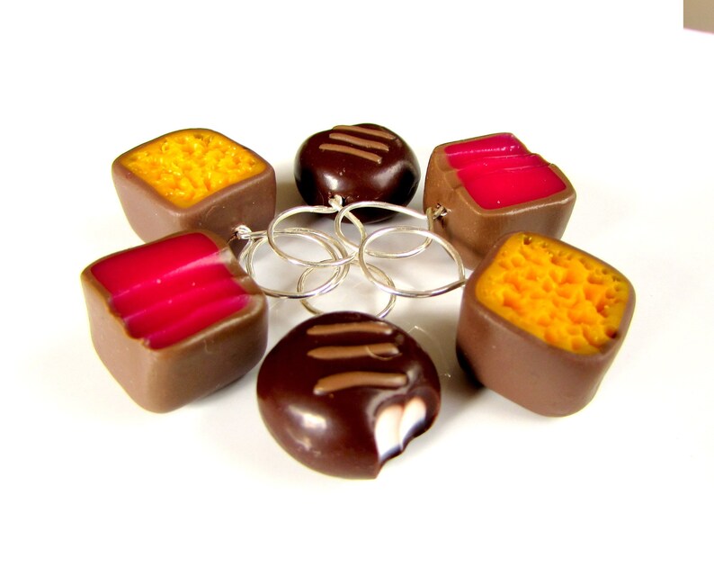 May include: Six chocolate candies with different fillings, including raspberry, orange, and chocolate. The candies are connected by silver rings.