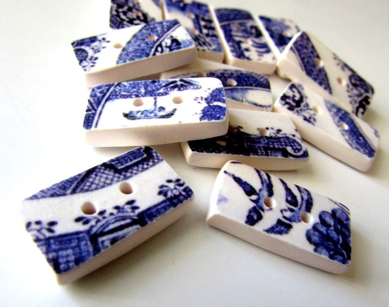 K&ouml;nnte beinhalten: Eine Sammlung rechteckiger Kn&ouml;pfe mit blau-wei&szlig;em Blumendesign. Jeder Knopf hat zwei L&ouml;cher und einen cremefarbenen Rand. Die Kn&ouml;pfe liegen auf einer wei&szlig;en Oberfl&auml;che.