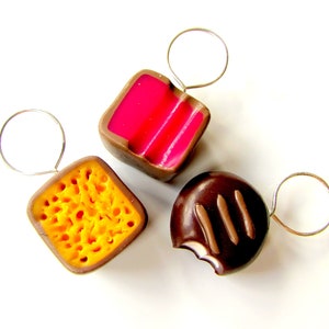 May include: Three miniature chocolate candies on silver rings. The candies are shaped like a square, a circle, and a honeycomb. The square candy is pink and white, the circle candy is brown with white stripes, and the honeycomb candy is yellow.