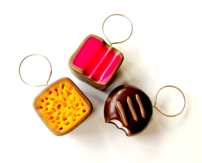 May include: Three miniature chocolate candies on silver rings. The candies are shaped like a square, a circle, and a honeycomb. The square candy is pink and white, the circle candy is brown with white stripes, and the honeycomb candy is yellow.