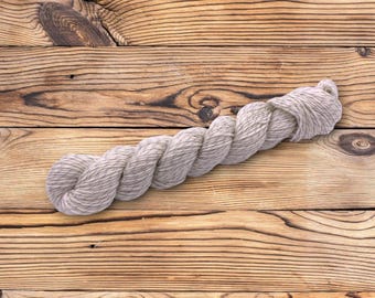 May include: A skein of light gray yarn, tightly twisted, lies on a wooden surface. The yarn appears to be made of a natural fiber, with subtle variations in color. The wooden background has a warm, natural tone with visible wood grain.