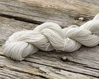 May include: A skein of white yarn with a subtle sheen, twisted and looped, resting on a weathered wooden surface.